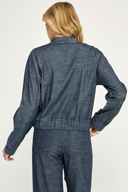 A woman shown from the back modeling a cropped, dark blue stargazer alex bomber denim jacket paired with matching denim pants against a light gray studio background.