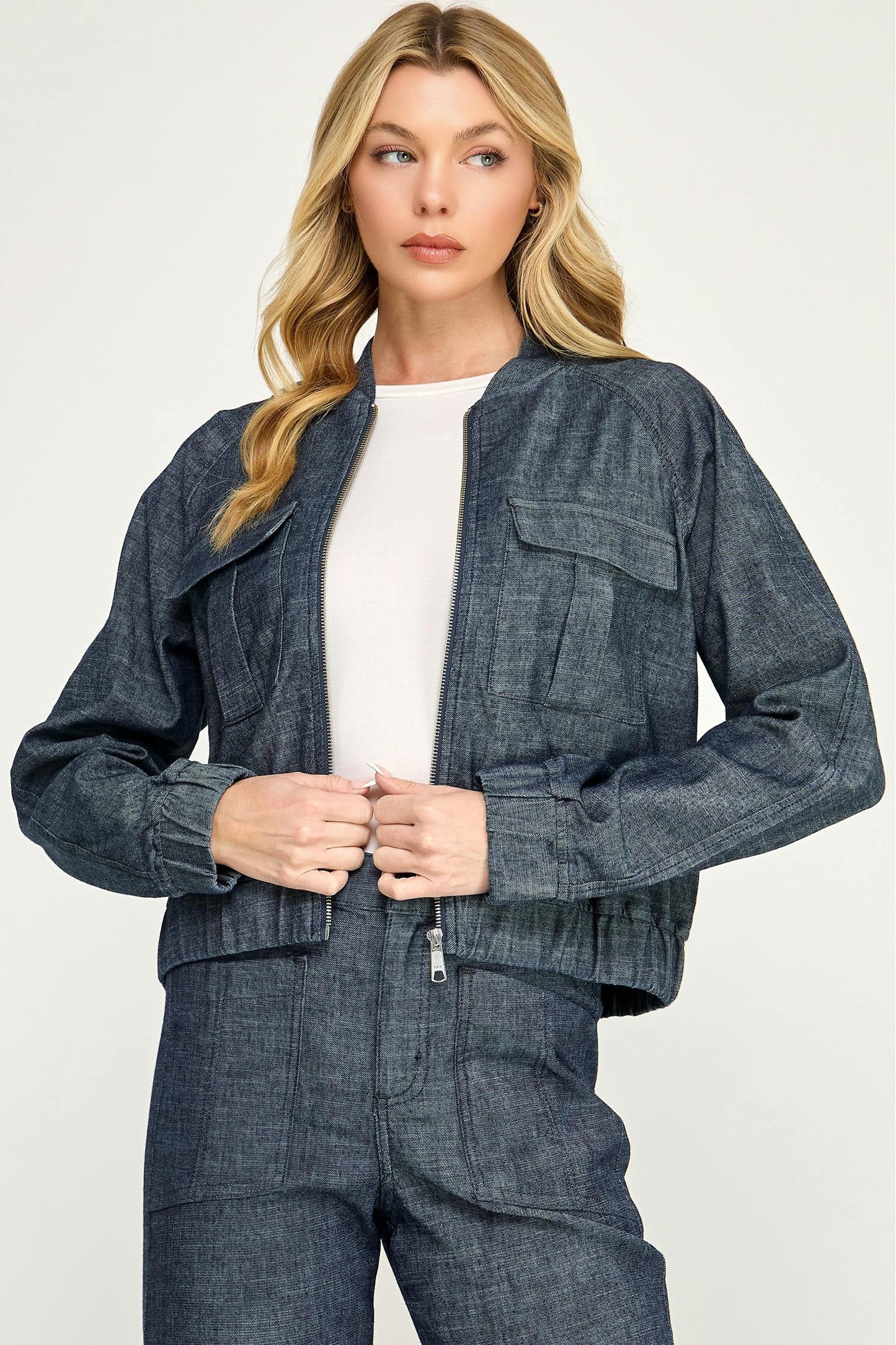 A woman modeling a cropped, dark blue  stargazer alex bomber denim jacket with a zip-up front, button closure, and flap chest pockets, paired with matching denim pants and a white top, against a light gray studio background.