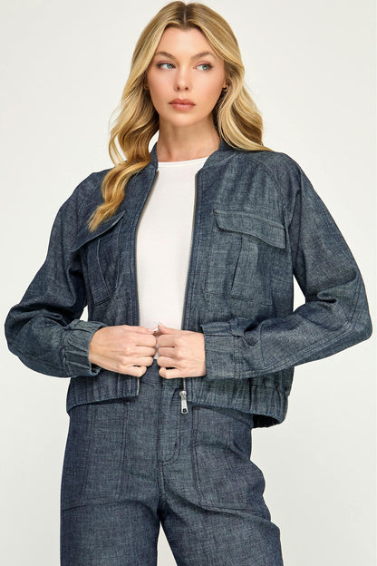 A woman modeling a cropped, dark blue  stargazer alex bomber denim jacket with a zip-up front, button closure, and flap chest pockets, paired with matching denim pants and a white top, against a light gray studio background.