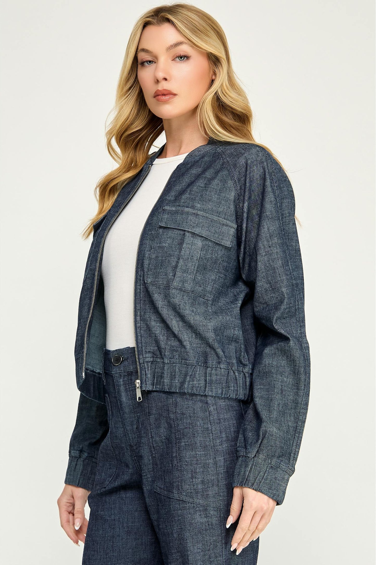 A woman modeling a cropped, dark blue  stargazer alex bomber denim jacket with a zip-up front, button closure, and flap chest pockets, paired with matching denim pants and a white top, against a light gray studio background.