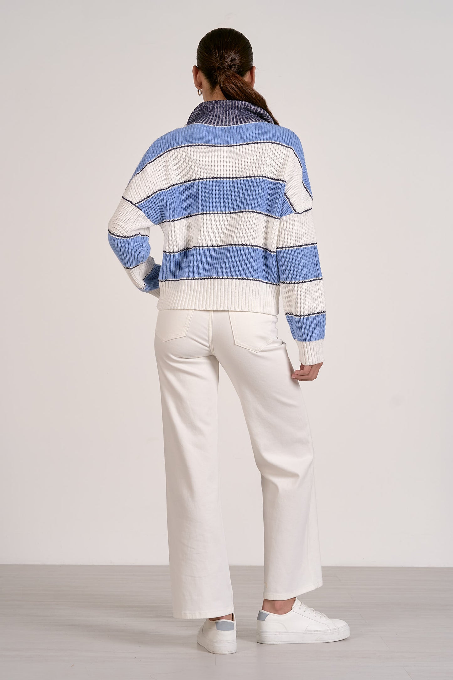 Back view of person wearing a light blue and white striped sweater with white pants on a plain background