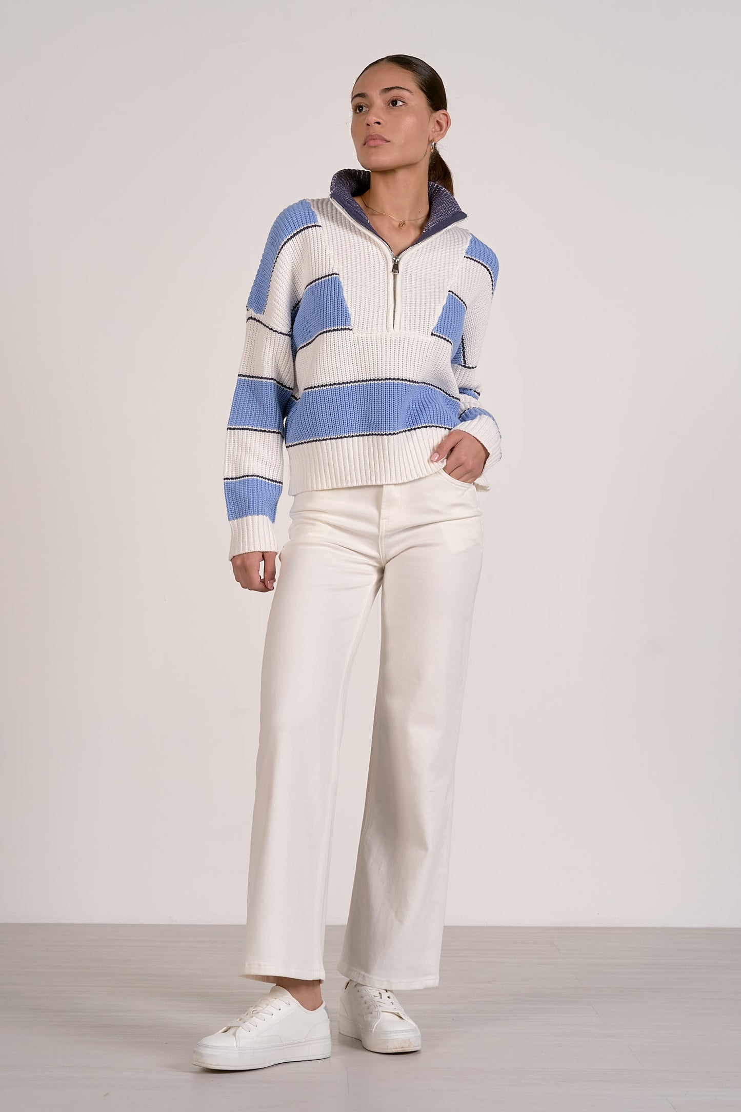 Person wearing a light blue and white striped quarter zip sweater and white pants on a plain background