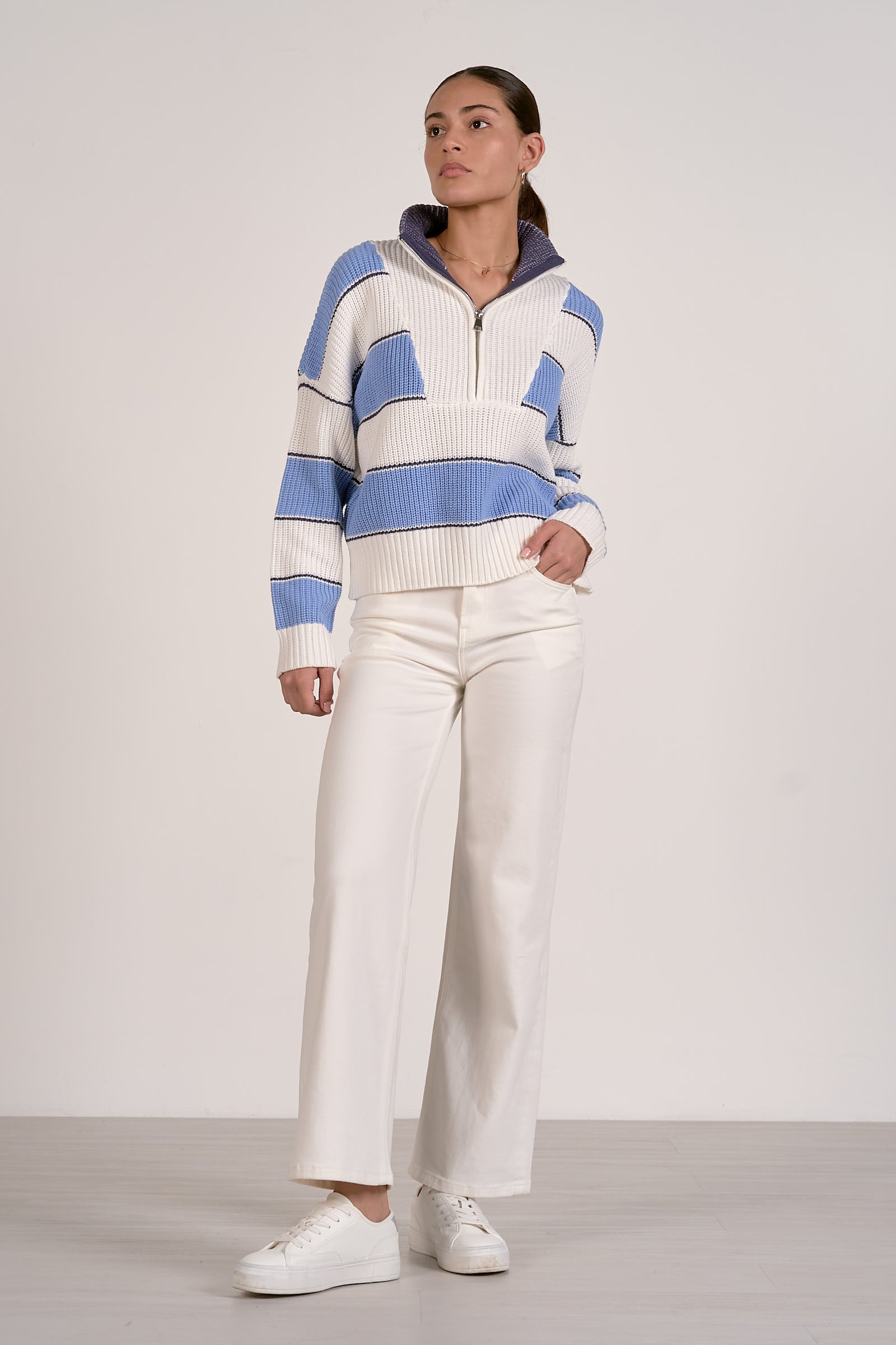 Person wearing a light blue and white striped quarter zip sweater and white pants on a plain background