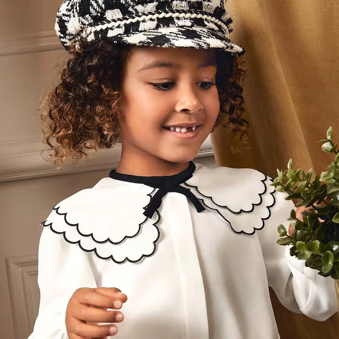 A young girl wears a white collared shirt and black and white houndstooth hat.