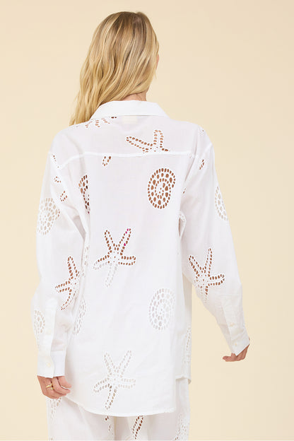 A woman shown from the back wearing a white long-sleeve button-up shirt with cutout starfish and circular seashell patterns, paired with matching white pants, against a solid beige background.