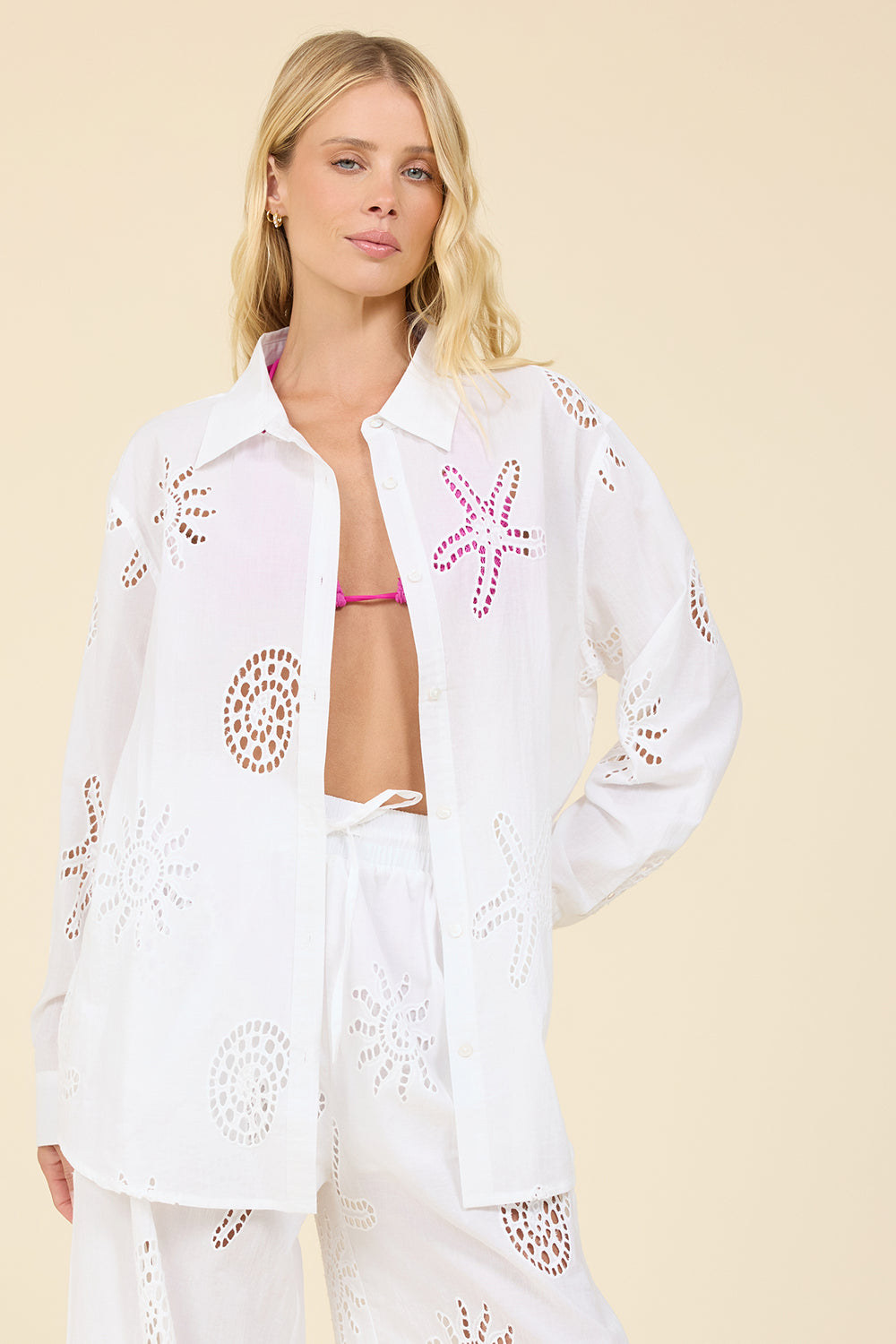 A woman modeling a white eyelet button-up shirt with pink starfish and brown circular cutout patterns, paired with matching white pants, against a solid beige background.