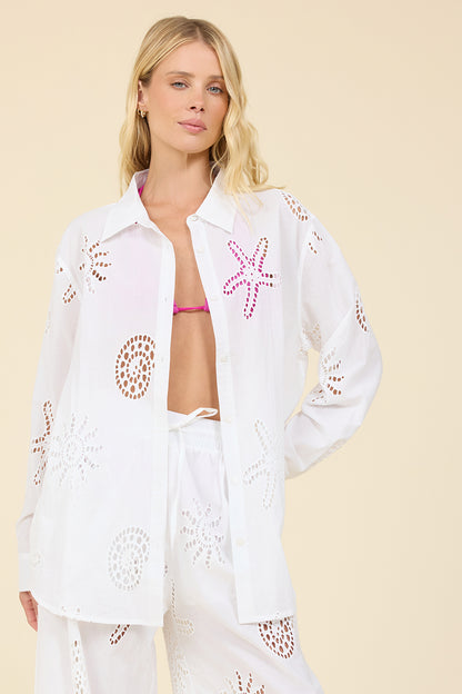 A woman modeling a white eyelet button-up shirt with pink starfish and brown circular cutout patterns, paired with matching white pants, against a solid beige background.