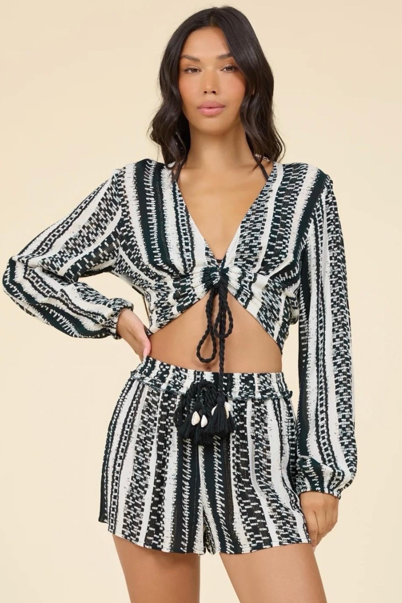 A woman modeling a matching black and white geometric patterned two-piece set consisting of a long-sleeve cropped tie-front top and high-waisted shorts, against a solid beige background.