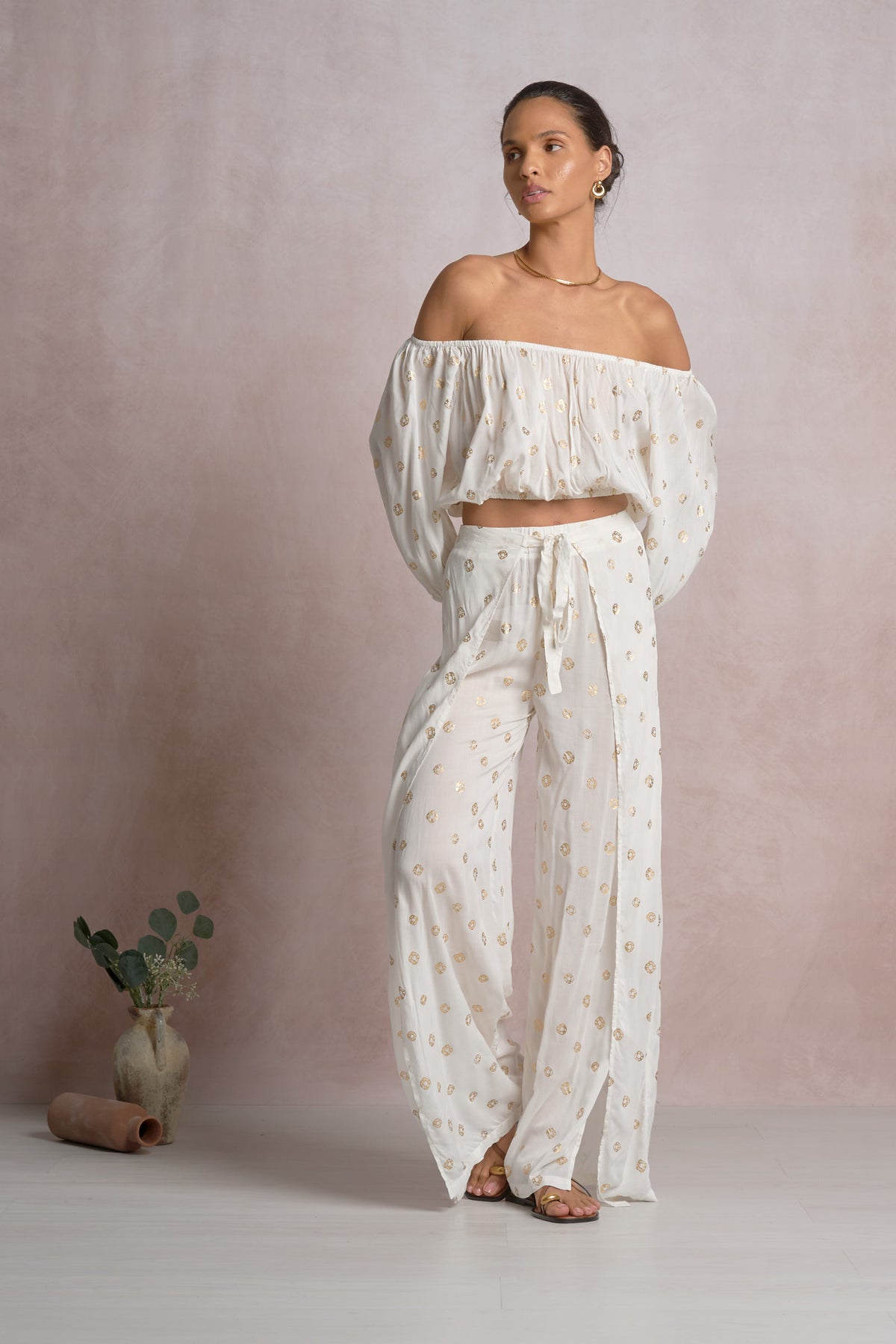Woman wearing a white off-shoulder top with gold polka dots and pants against a plain background