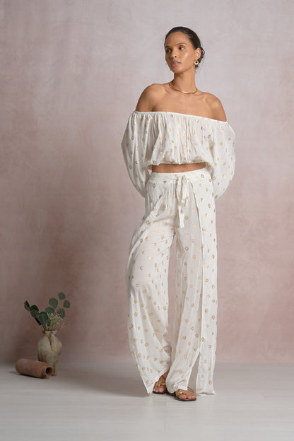 Woman wearing a white off-shoulder top with gold polka dots and pants against a plain background