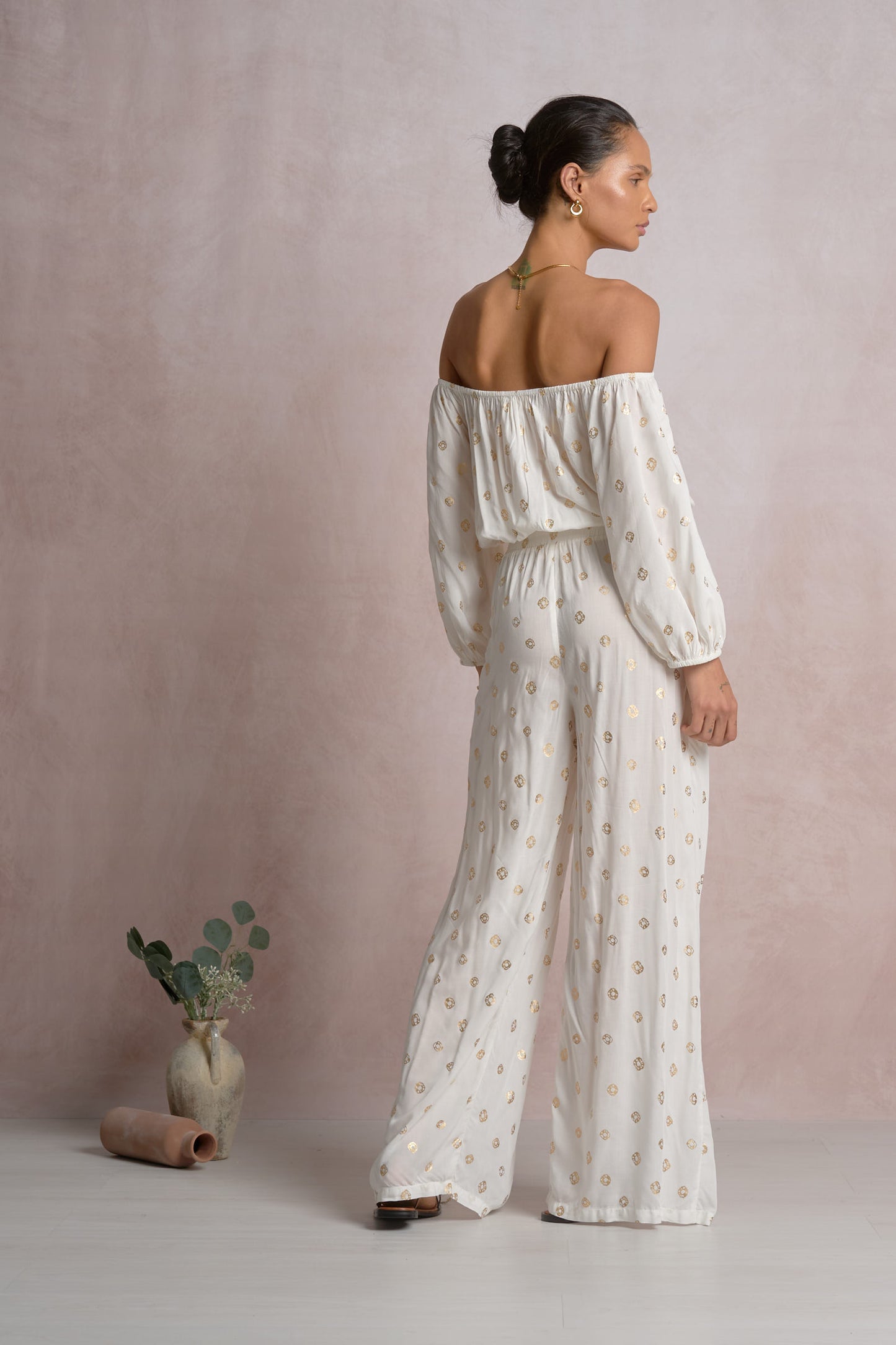 Woman wearing a white off-shoulder top with gold polka dots and wide-leg pants against a plain background
