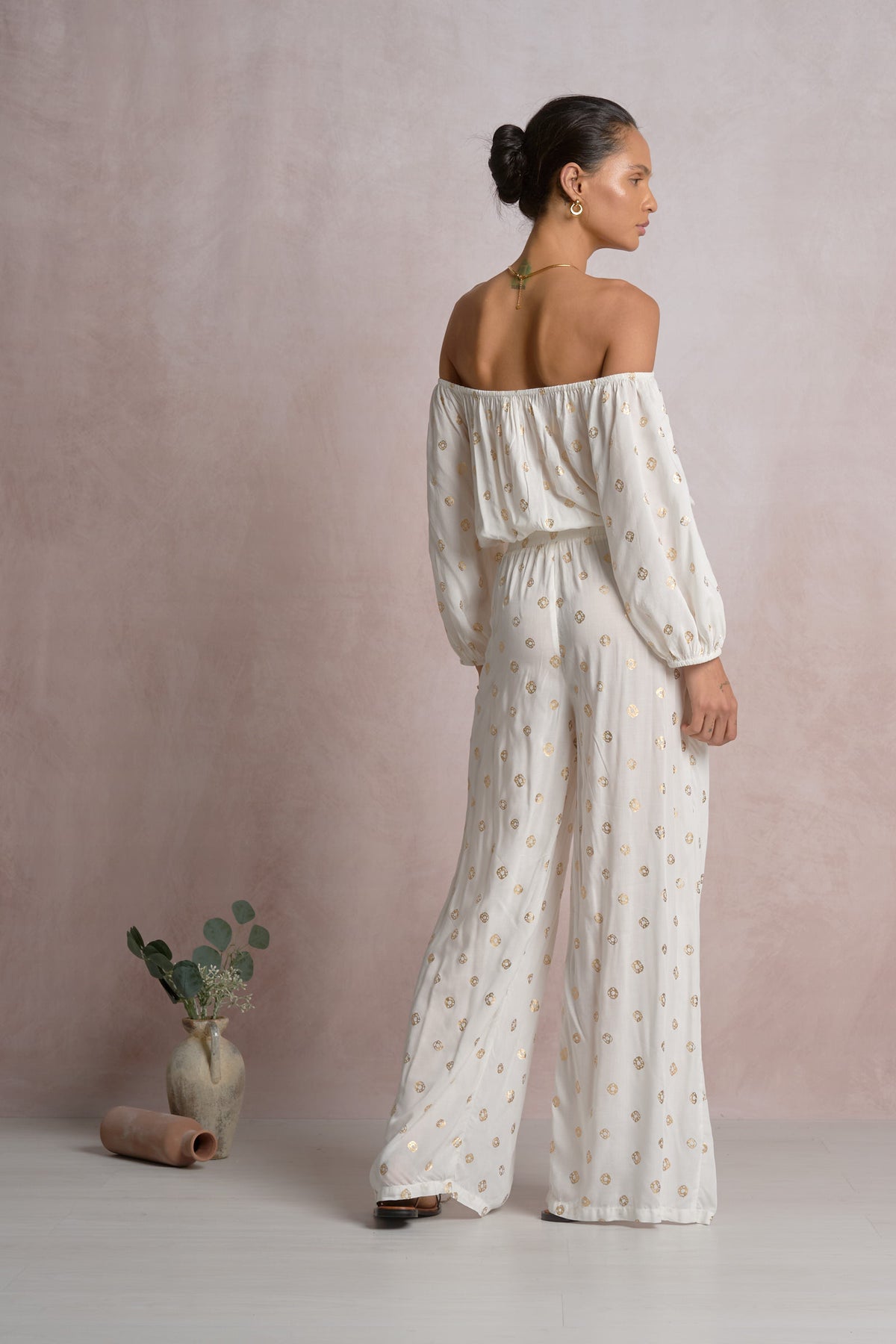 Woman wearing a white off-shoulder top with gold polka dots and wide-leg pants against a plain background