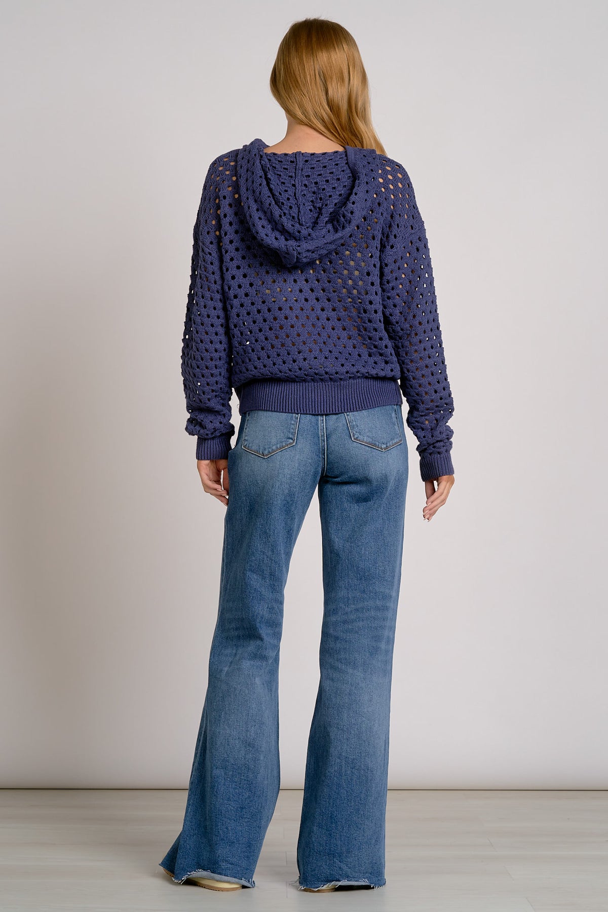 A woman viewed from the back wearing a navy blue open-knit cropped hoodie and blue flare jeans, standing against a plain light gray studio background.