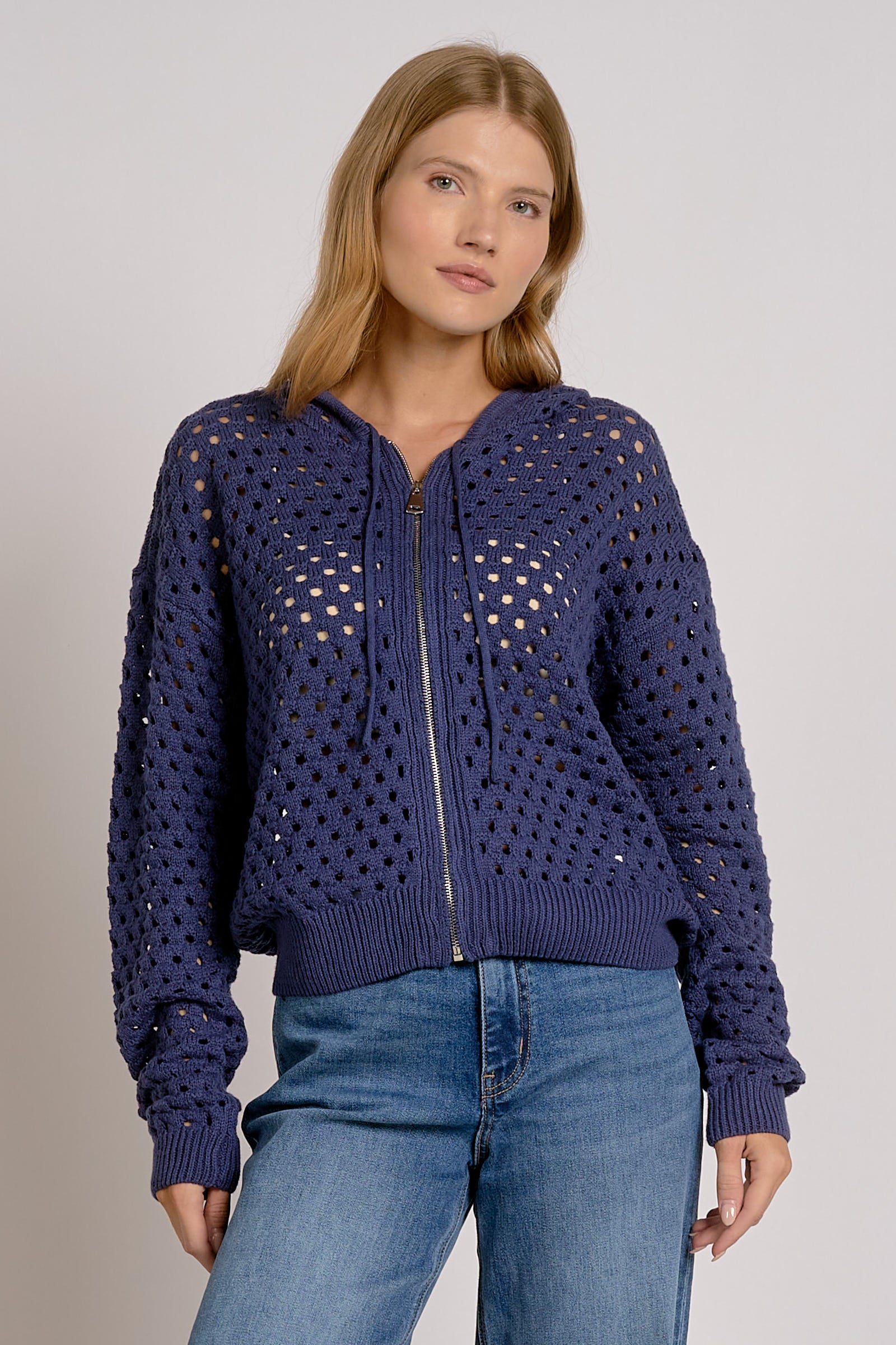 A woman wearing a navy blue open-knit zip-up hoodie with a ribbed hem and cuffs, paired with blue jeans, against a light gray studio background.
