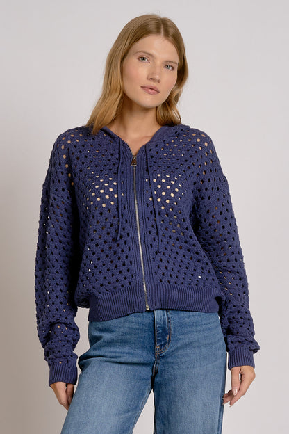 A woman wearing a navy blue open-knit zip-up hoodie with a ribbed hem and cuffs, paired with blue jeans, against a light gray studio background.