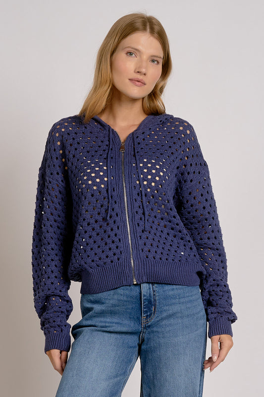 A woman wearing a navy blue open-knit zip-up hoodie with a ribbed hem and cuffs, paired with blue jeans, against a light gray studio background.