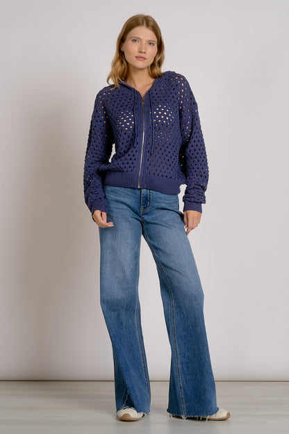 A woman modeling a navy blue open-knit zip-up hoodie with wide-leg blue jeans, standing against a neutral light gray studio background.