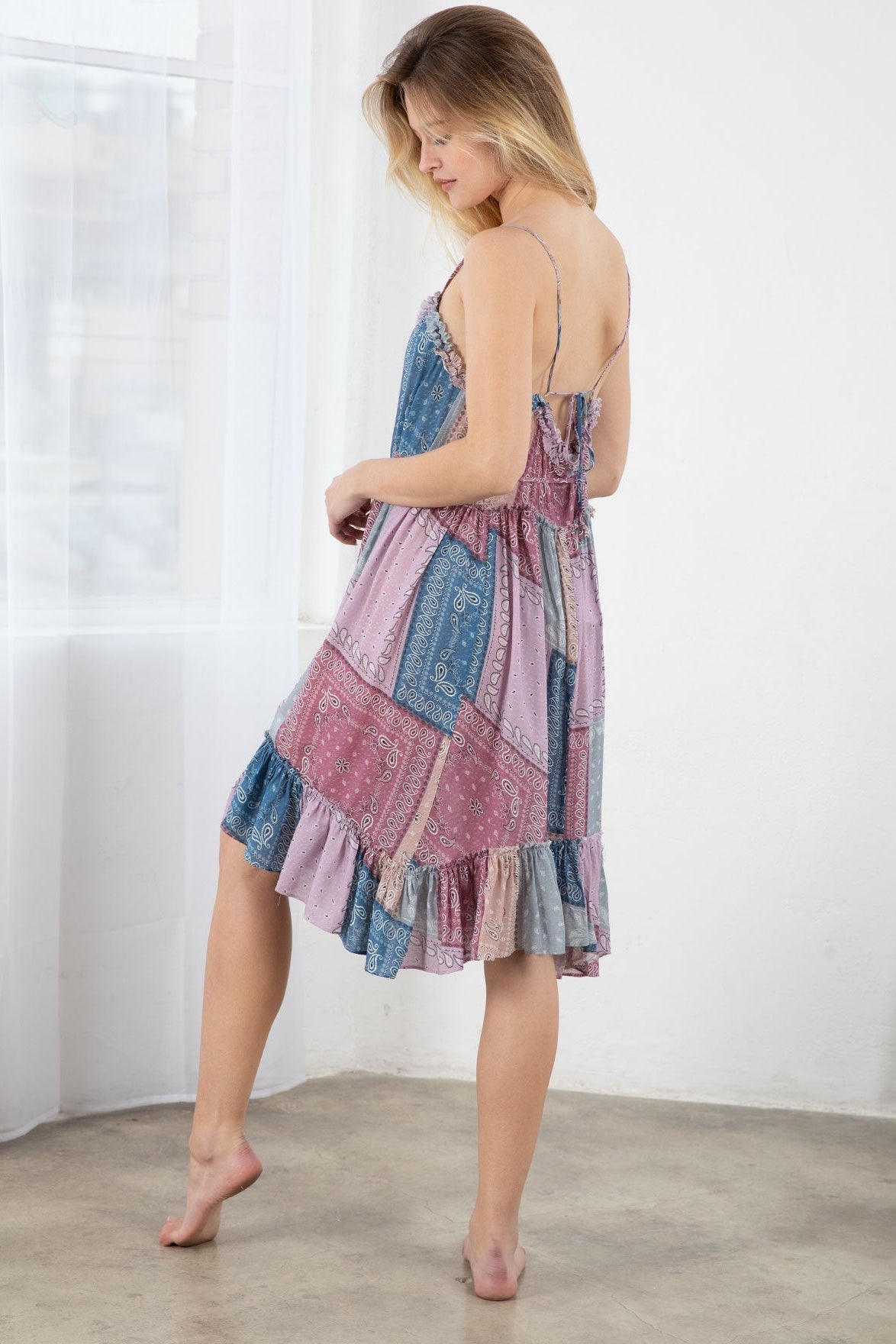 A woman modeling a bohemian patchwork print midi dress with lavender blue denim and pink, purple, and beige patterned panels, shown from the back in a bright room with white curtains.