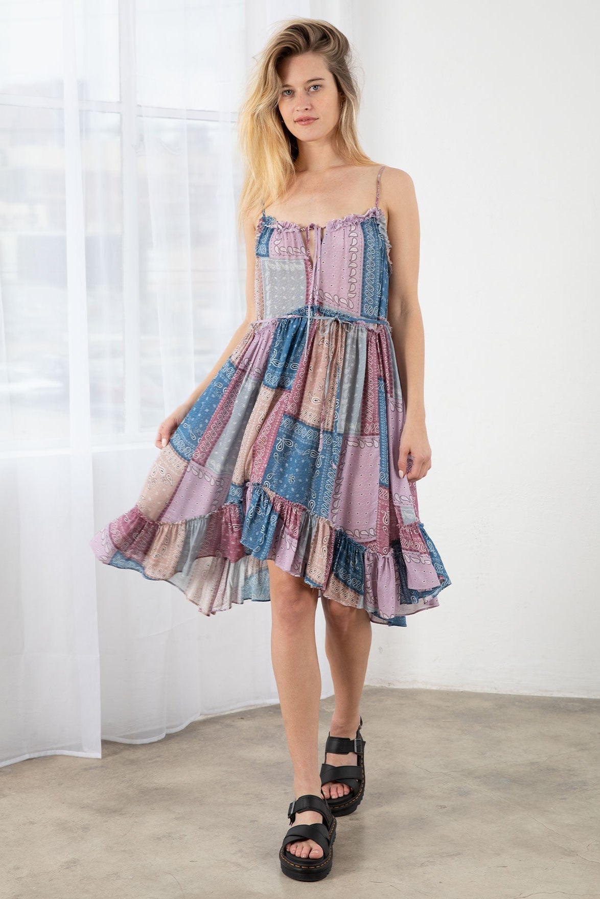 A woman modeling a tiered lavender blue patchwork mini dress with spaghetti straps and a split neckline, featuring a multicolor print of dusty blue, pink, purple, and beige. She is wearing black platform sandals in a bright, minimalist room with white curtains and a concrete floor.