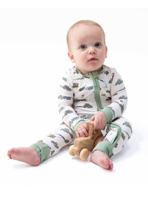 A baby sitting on a white studio background wearing a long-sleeve footless pajama romper with a green and brown farm machines print, holding a small wooden toy car.