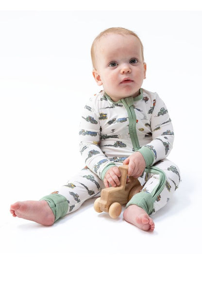A baby sitting on a white studio background wearing a long-sleeve footless pajama romper with a green and brown farm machines print, holding a small wooden toy car.