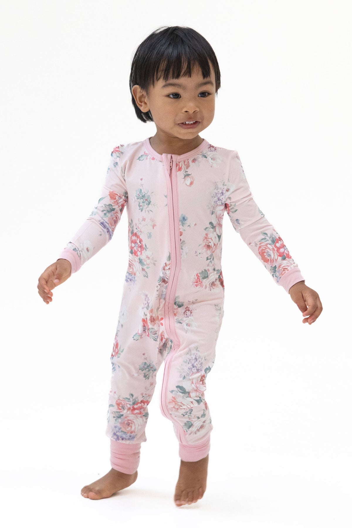 A toddler standing barefoot against a plain white studio background, wearing a light pink painterly rose long-sleeve footless one-piece pajama romper with a front zipper.