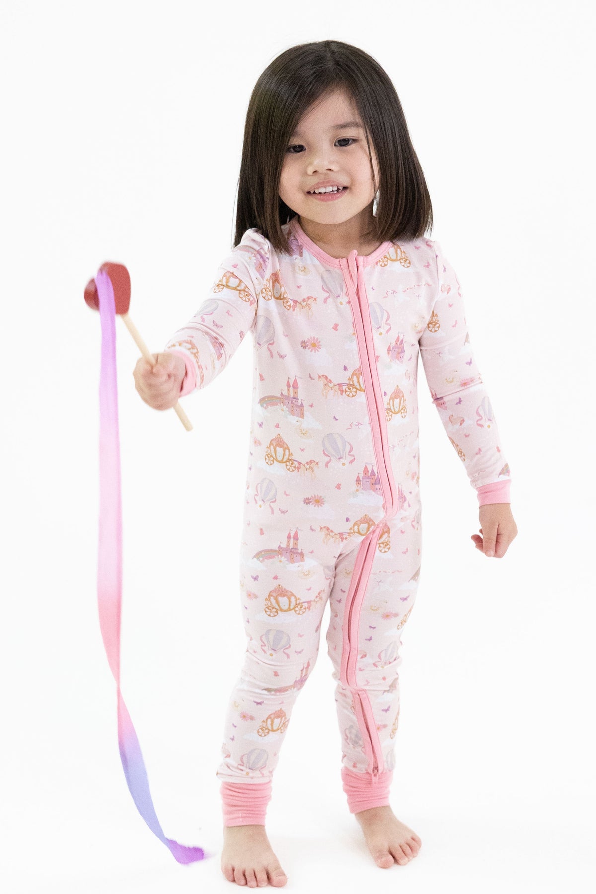 A young child wearing a pink one-piece romper pajama with a princess unicorn print stands barefoot against a white background, holding a pink and purple ribbon wand.