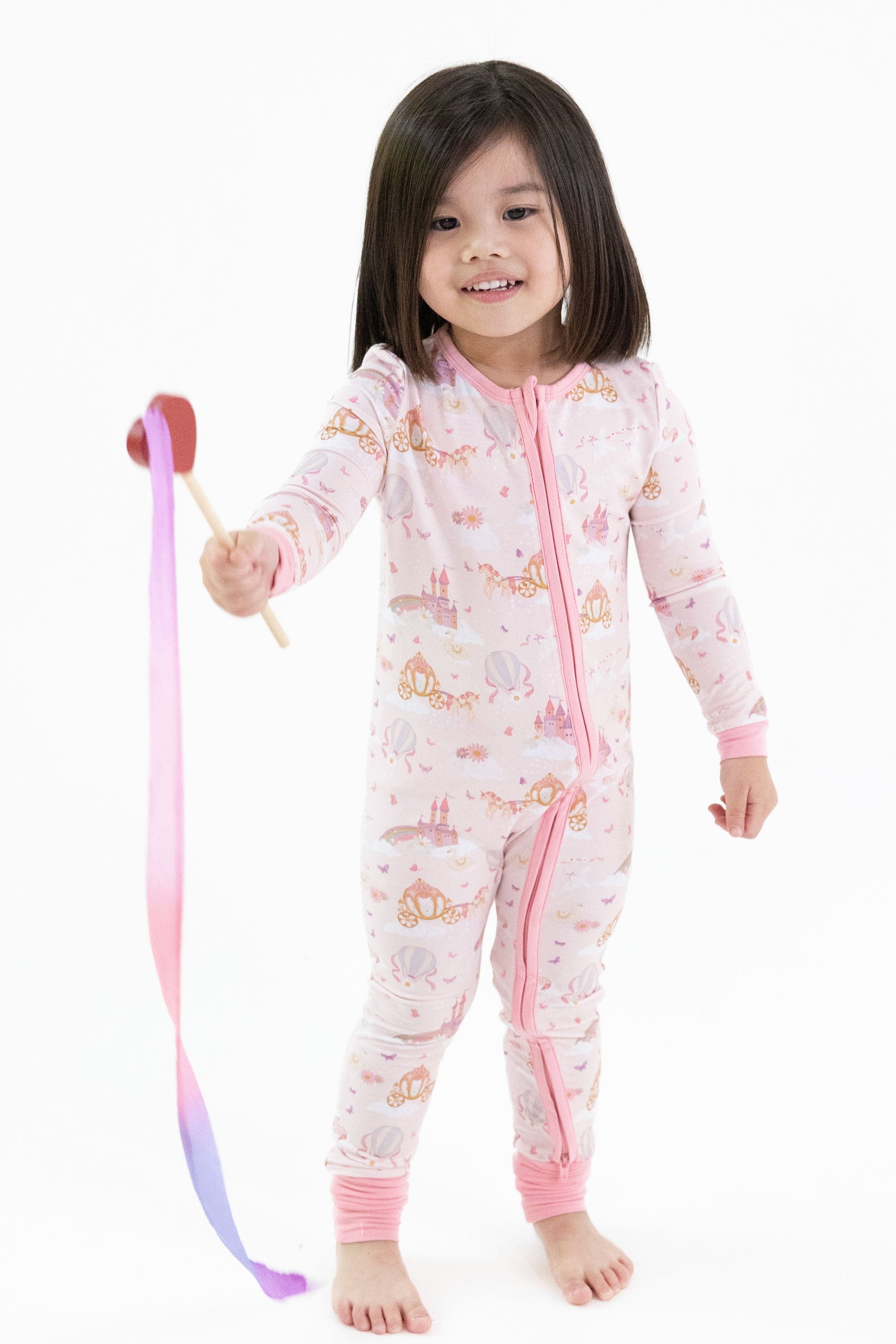 A young child wearing a pink one-piece romper pajama with a princess unicorn print stands barefoot against a white background, holding a pink and purple ribbon wand.
