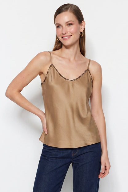 A woman modeling a satin-like taupe camisole top with thin straps and decorative trim along the neckline, paired with dark blue jeans against a plain light gray studio background.