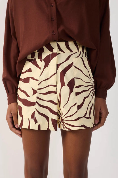 A person wearing high-waisted shorts with a bold brown and cream abstract alvorecer pattern, paired with a brown button-up blouse, shown from the chest down against a light gray studio background.