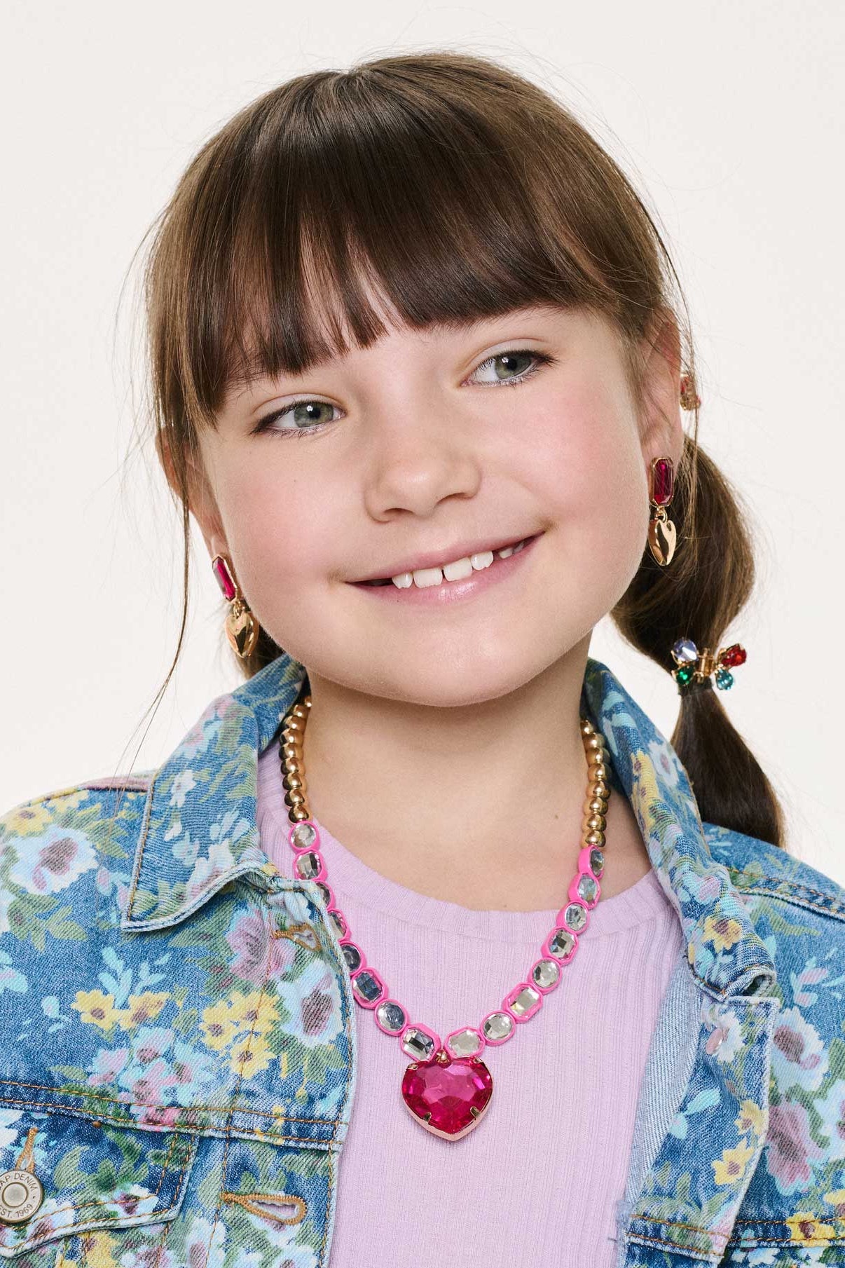 Young girl wearing a floral denim jacket and pink mega set jewelry against a light background