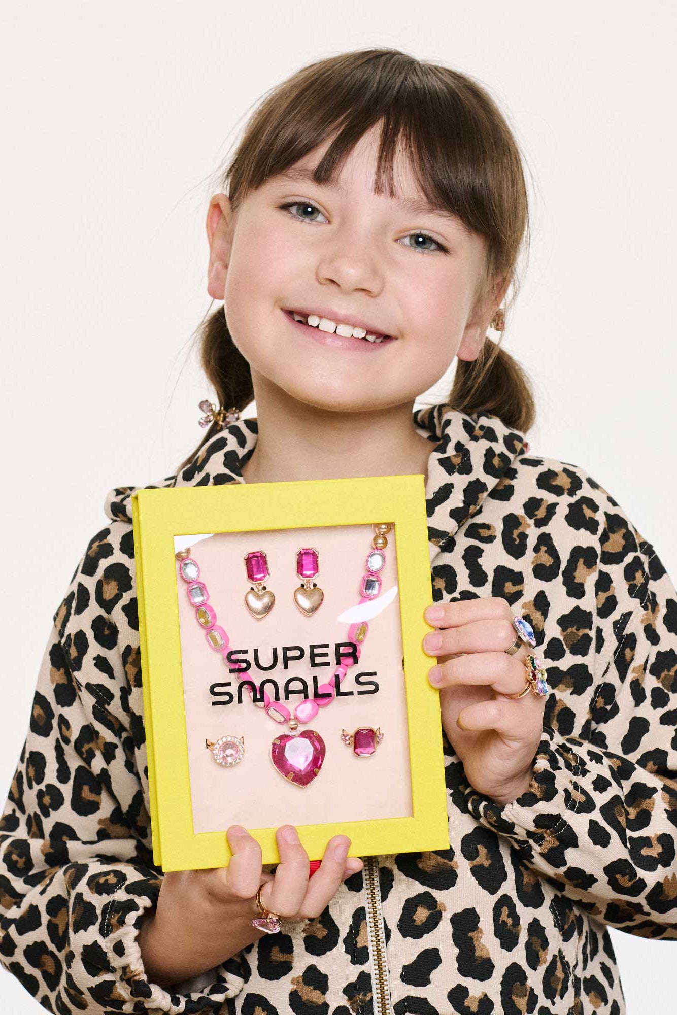 Child holding a yellow frame with pink jewelry and 'Super Smalls' text against a plain background