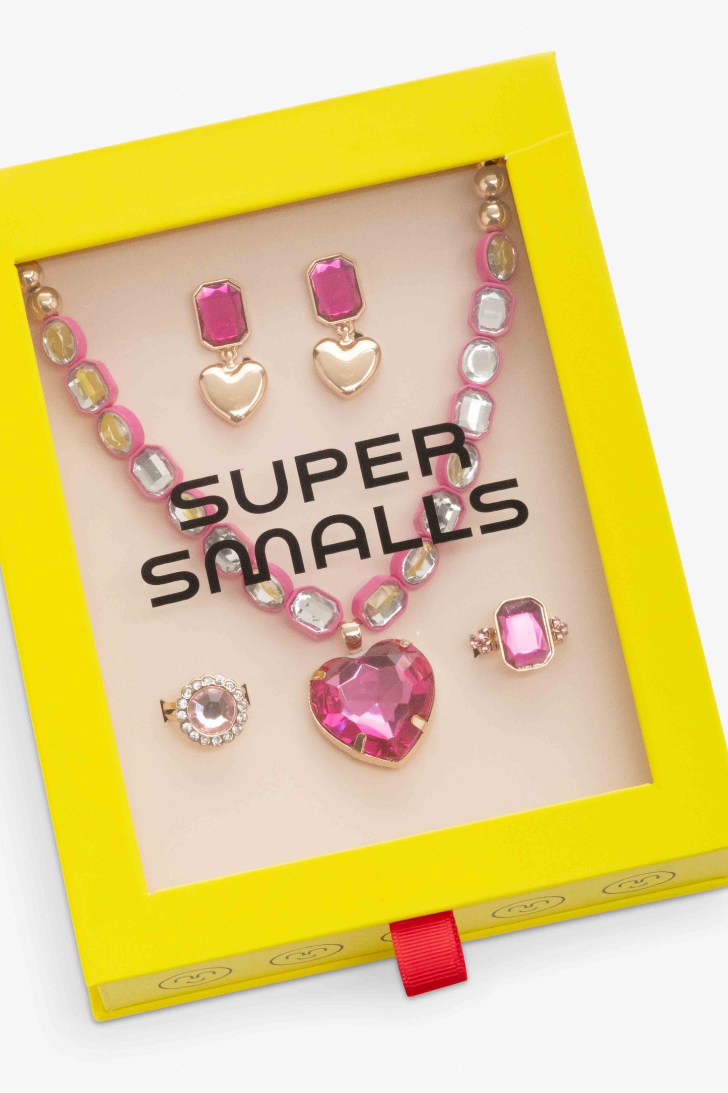 A jewelry set displayed inside a bright yellow 'Super Smalls' gift box with a clear window. The set includes a pink heart pendant necklace, matching heart and rectangular stud earrings, a ring, and a brooch, all with pink and gold details.