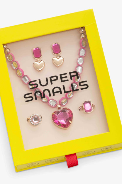 A jewelry set displayed inside a bright yellow 'Super Smalls' gift box with a clear window. The set includes a pink heart pendant necklace, matching heart and rectangular stud earrings, a ring, and a brooch, all with pink and gold details.