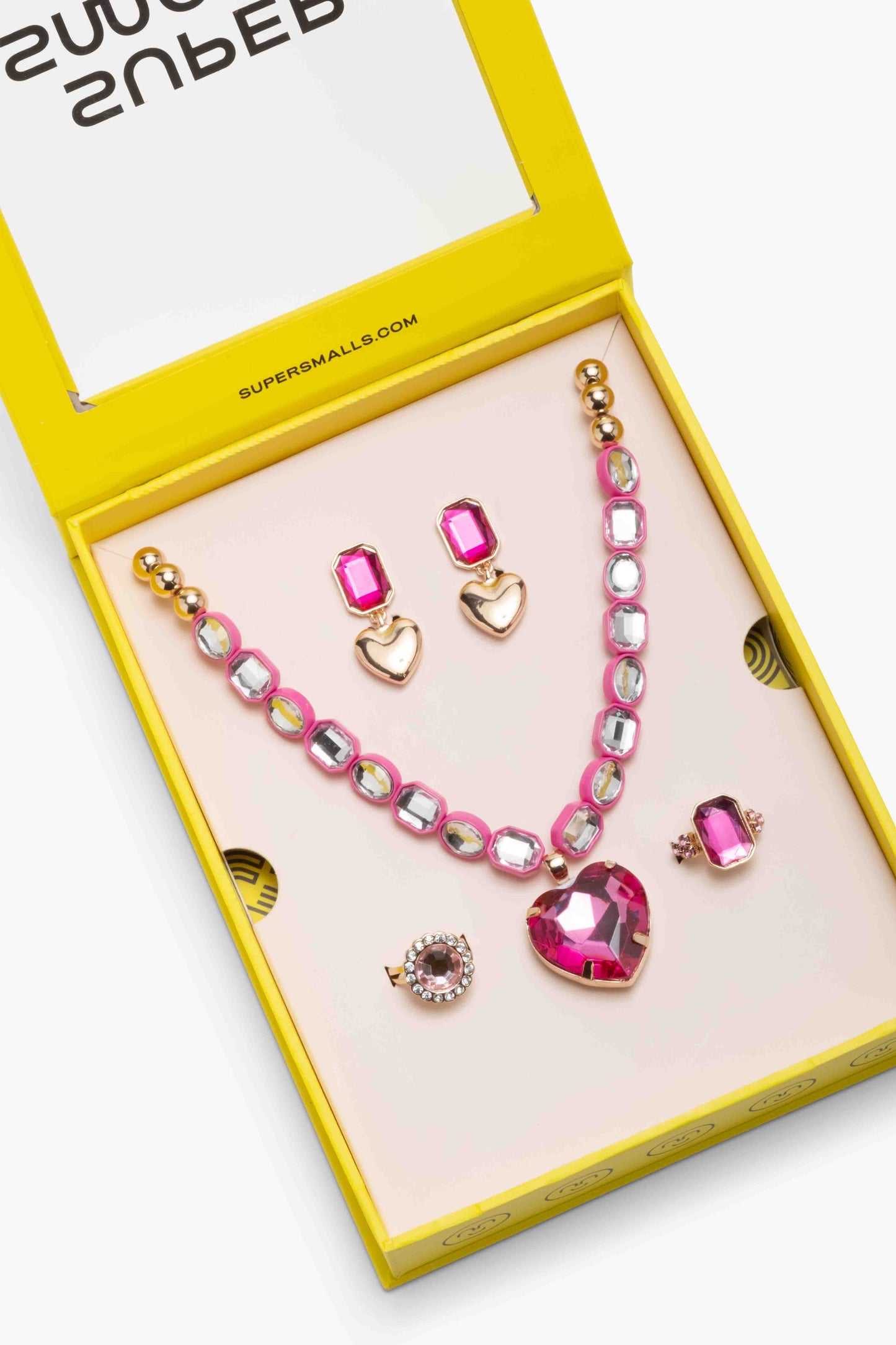 A pink and gold jewelry set with a necklace, earrings, and rings displayed inside a bright yellow Super Smalls box on a white background.