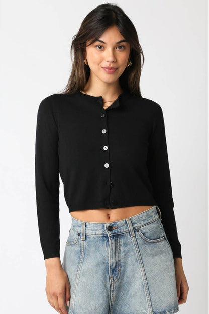 A woman wearing a black long-sleeve cropped button-front cardigan top and light-wash blue denim skirt, standing against a plain light gray studio background.