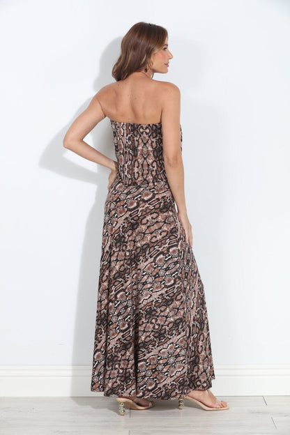 A woman modeling a strapless maxi dress with a brown, black, and beige snake print pattern, shown from the back against a white wall.