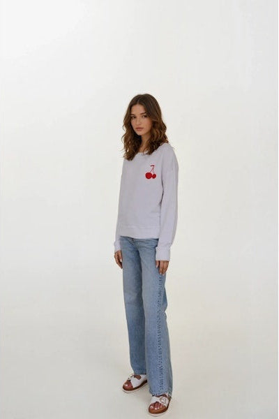 Woman wearing a white 
sweatshirt with a cherry design and blue jeans on a white background