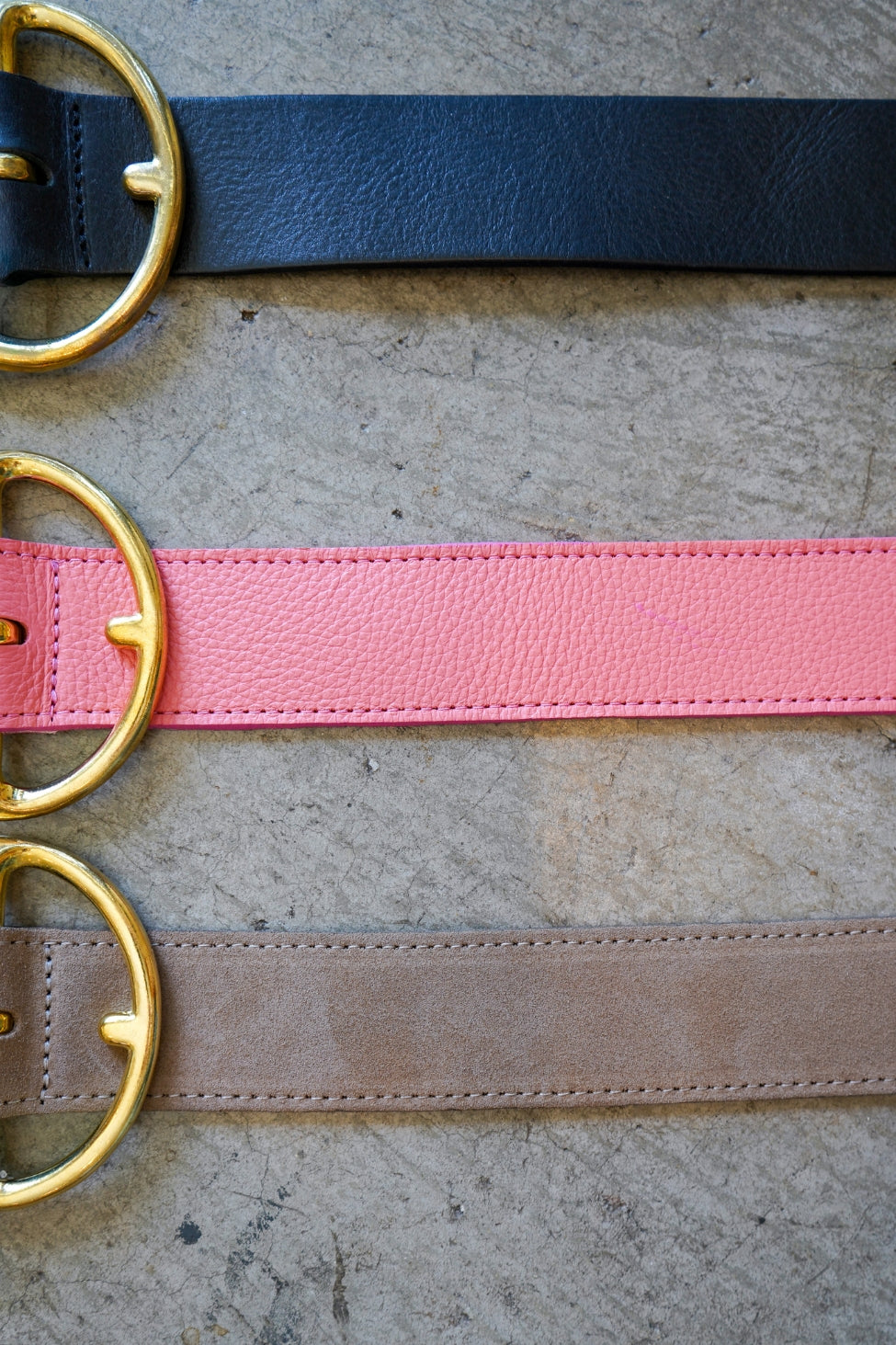 Three belts with gold buckles in black, pink, and brown on a textured surface.