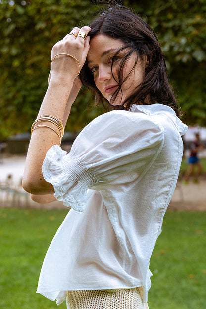 Woman in a white blouse standing outdoors with greenery in the background