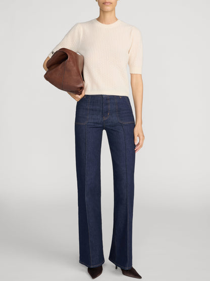 A model wearing a cream short-sleeve ribbed knit top, le slim palazzo pintuck pocket jeans in color Rinse, and dark brown pointed-toe heels, holding a brown leather clutch against a light gray studio background.