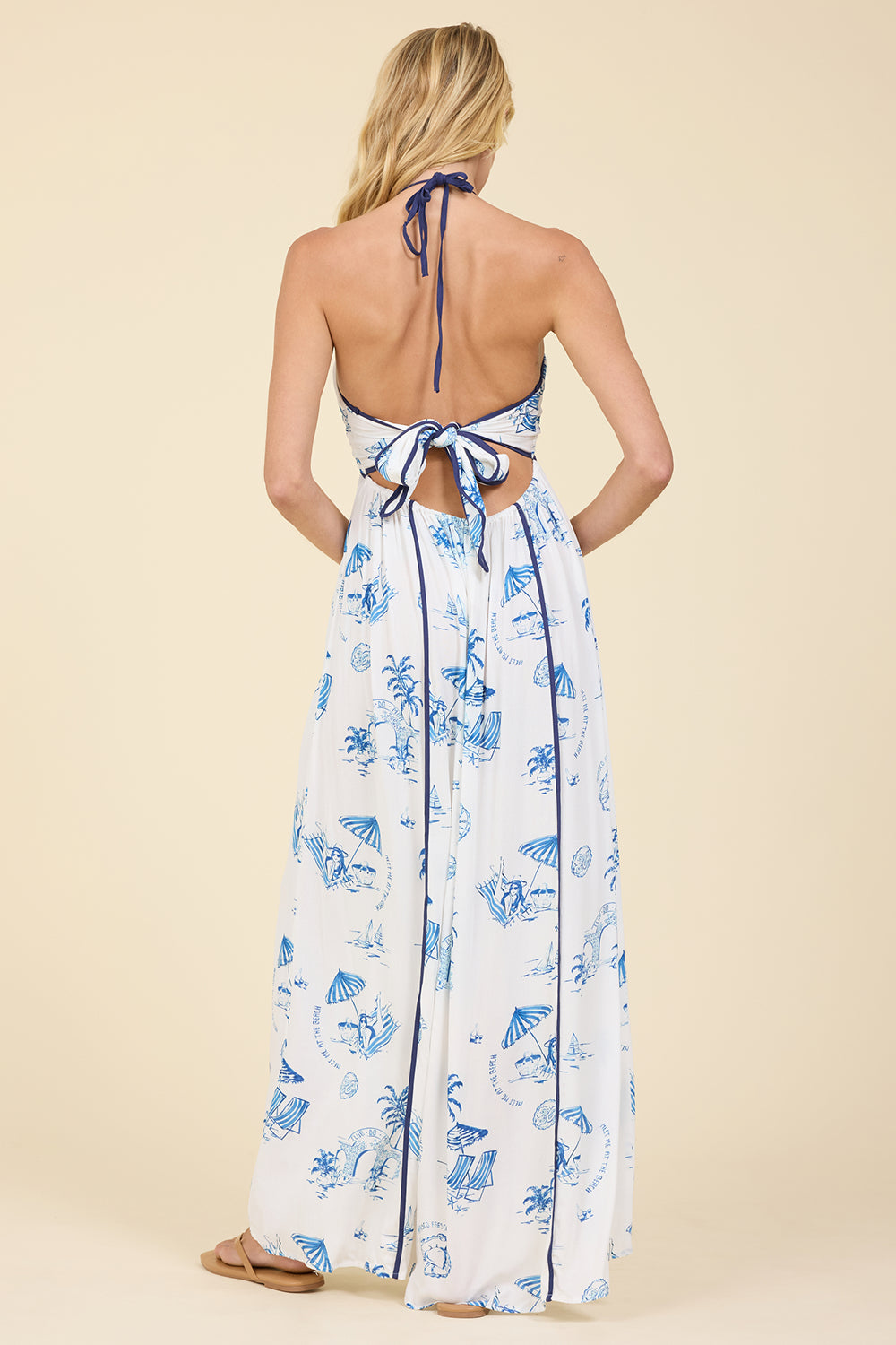 A woman shown from the back wearing a white halter maxi dress with a blue tropical "meet me at the beach" print and tie-back detail, standing against a solid beige studio background.
