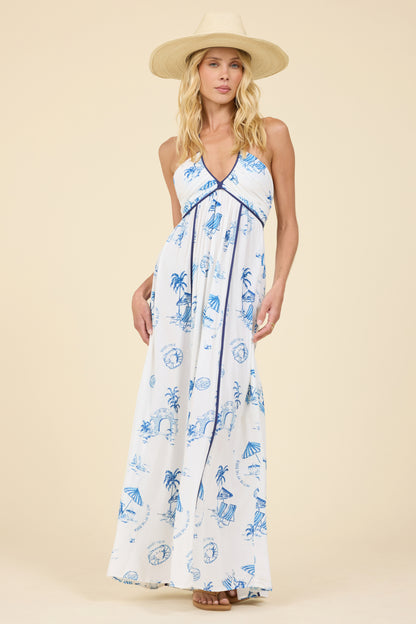 A woman modeling a white halter maxi dress with a blue tropical beach-themed  "Meet me at the beach" print, wearing a wide-brimmed straw hat against a solid beige studio background.