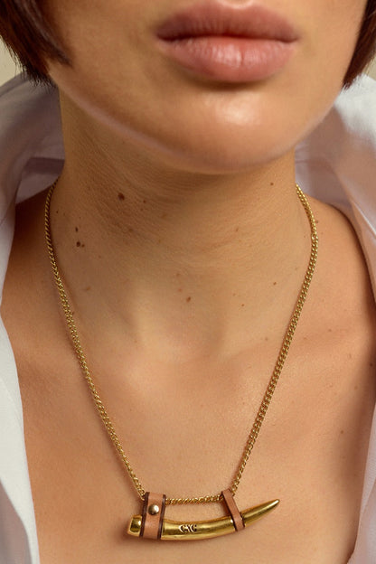 A close-up of a model wearing a gold chain necklace with a unique pendant featuring a gold fang element and a small brown leather strap detail.