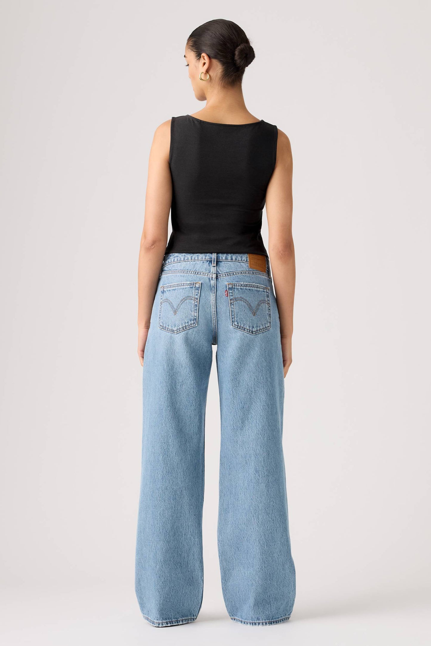 A model shown from the back wearing light-wash shakey ground wide-leg denim jeans and a black sleeveless top, standing against a plain light gray studio background.