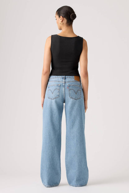 A model shown from the back wearing light-wash shakey ground wide-leg denim jeans and a black sleeveless top, standing against a plain light gray studio background.