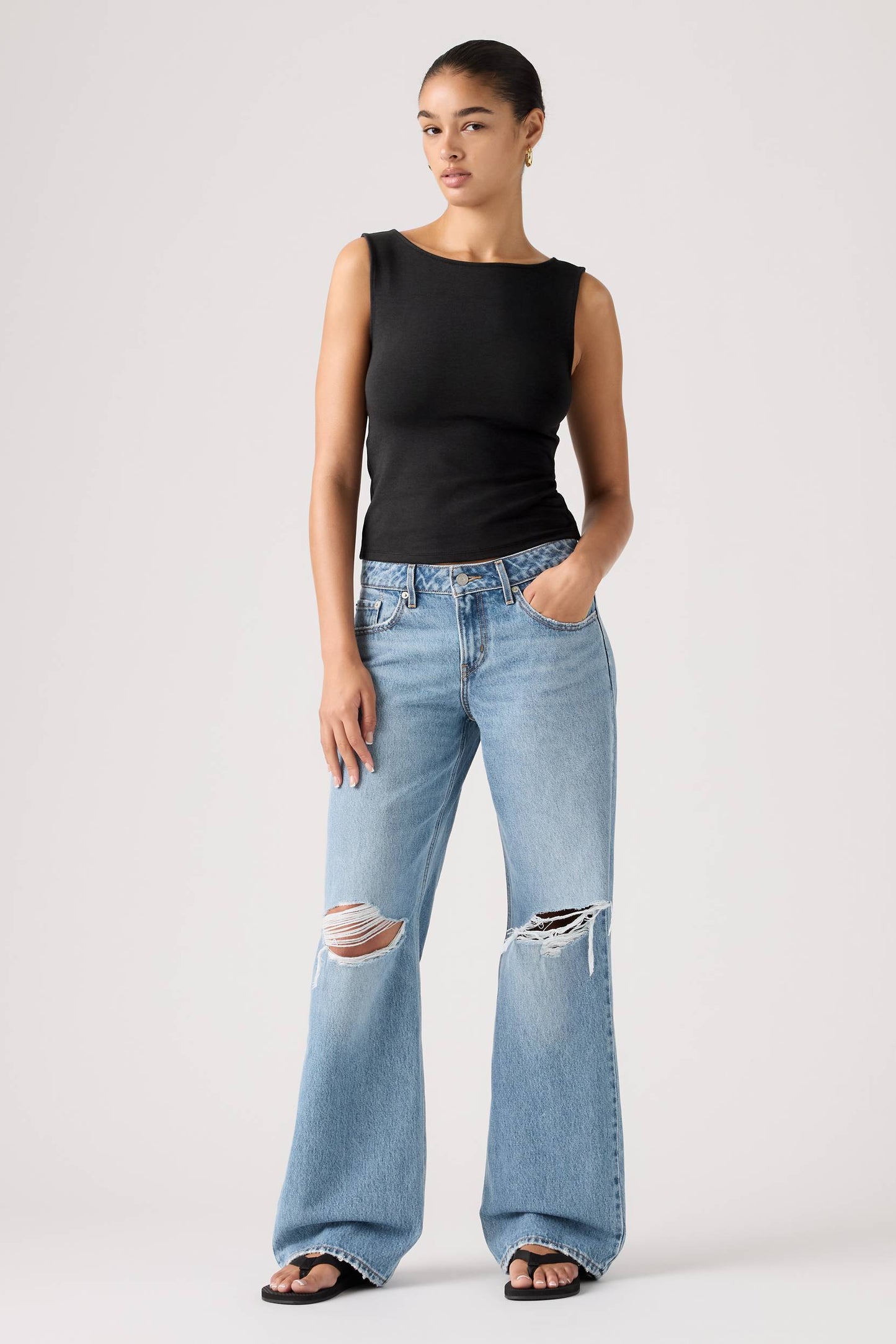 A model stands against a light gray studio background wearing a black sleeveless top and light-wash shakey ground wide-leg distressed jeans with ripped knees.