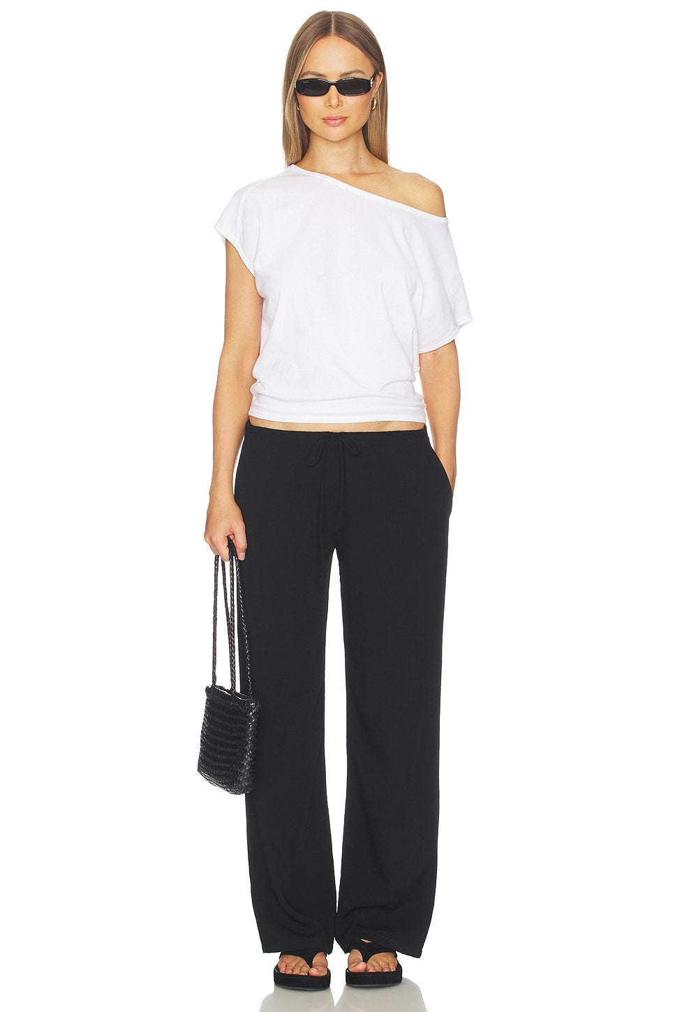 Woman wearing a white off-shoulder top and black pants, holding a black handbag on a white background.