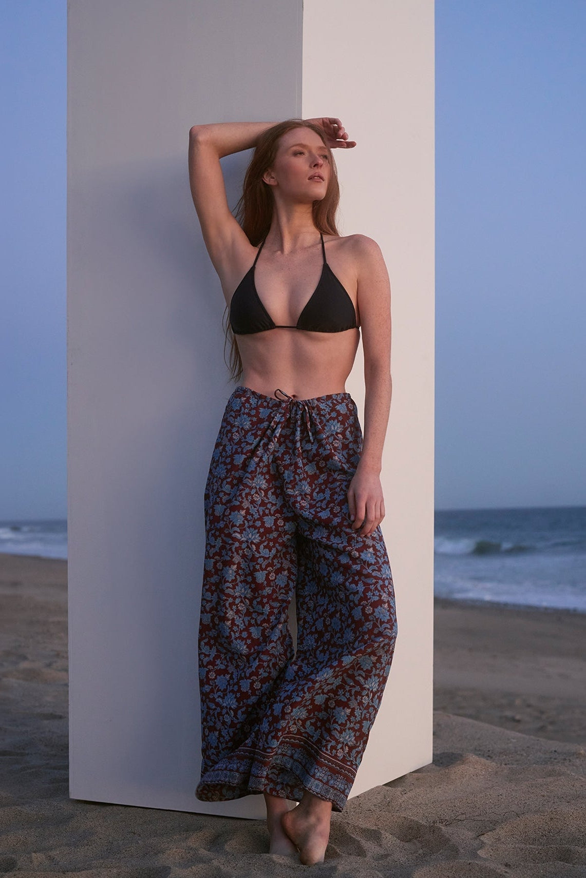 Woman in a black bikini top and patterned pants standing on a beach.