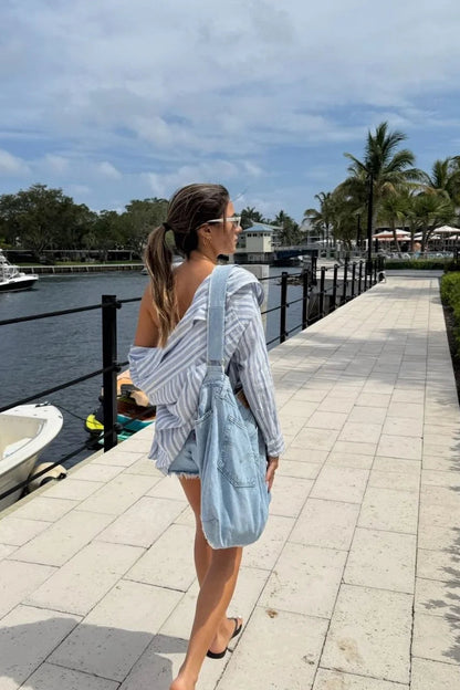 Woman walking along a waterfront path with boats and palm trees in the background carrying a denim purse 